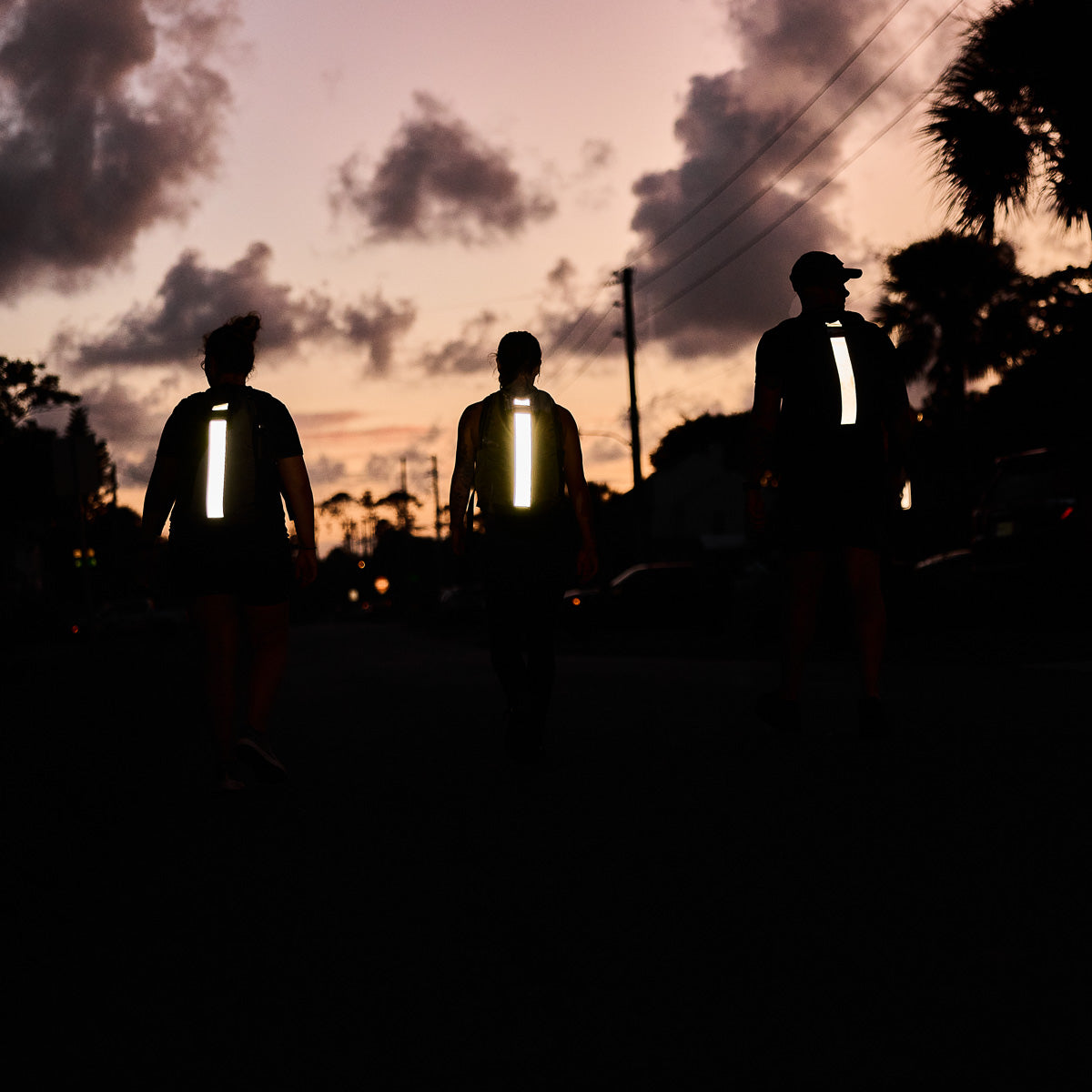 Three people with reflective strips on their Basic Rucker® backpacks walk at dusk beneath a cloudy sky and silhouetted palm trees.