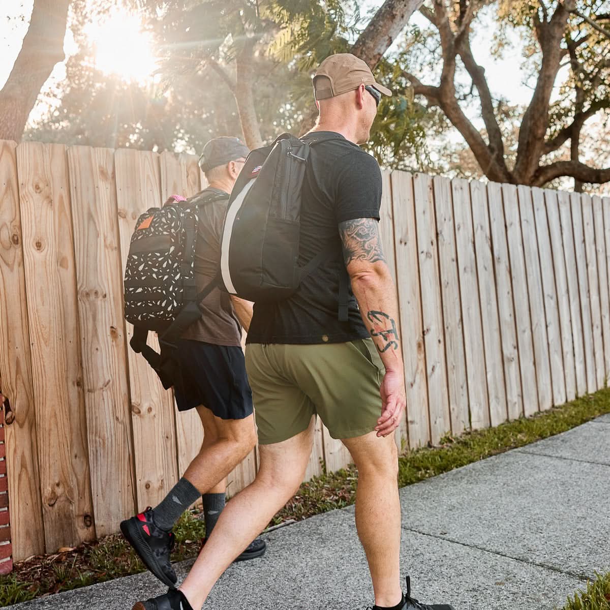 Two men with backpacks, including a Basic Rucker®, walk on a sidewalk next to a wooden fence on a sunny day.