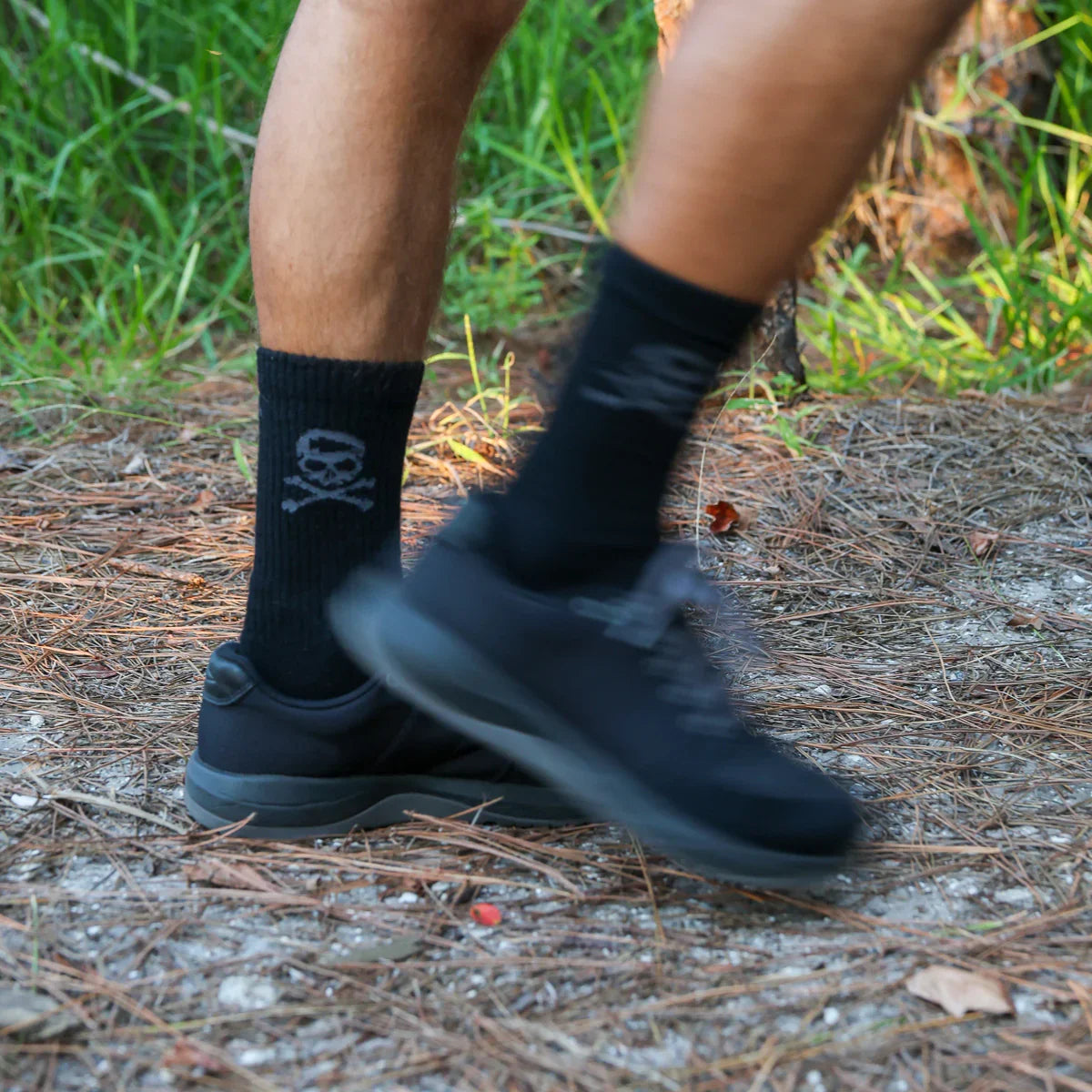 Close-up of a person wearing black GORUCK socks with skull logo and black shoes outdoors on a forest ground