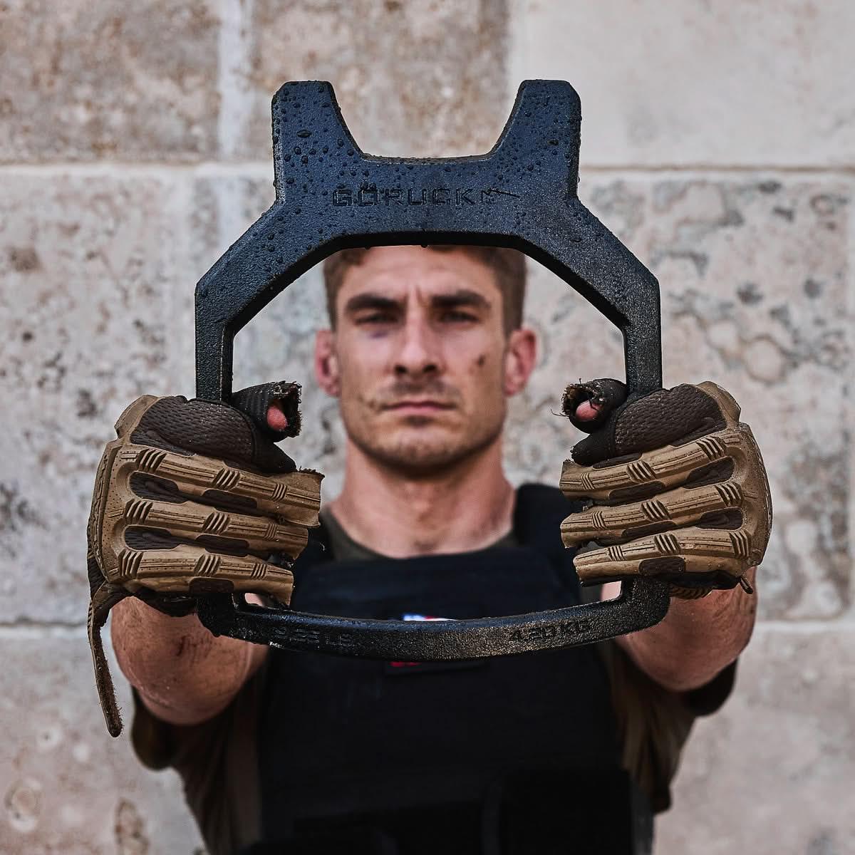 Man wearing tactical gloves and vest holding GORUCK steel training weight outdoors