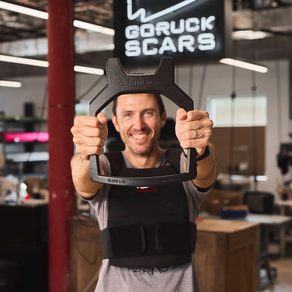 Man holding GORUCK ruck weight, wearing training vest in workshop with GORUCK SCARS sign