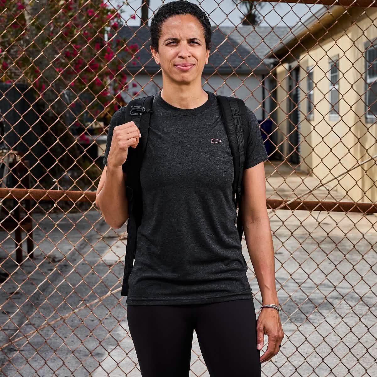 Person in black GORUCK t-shirt and backpack standing in front of chain-link fence outdoors