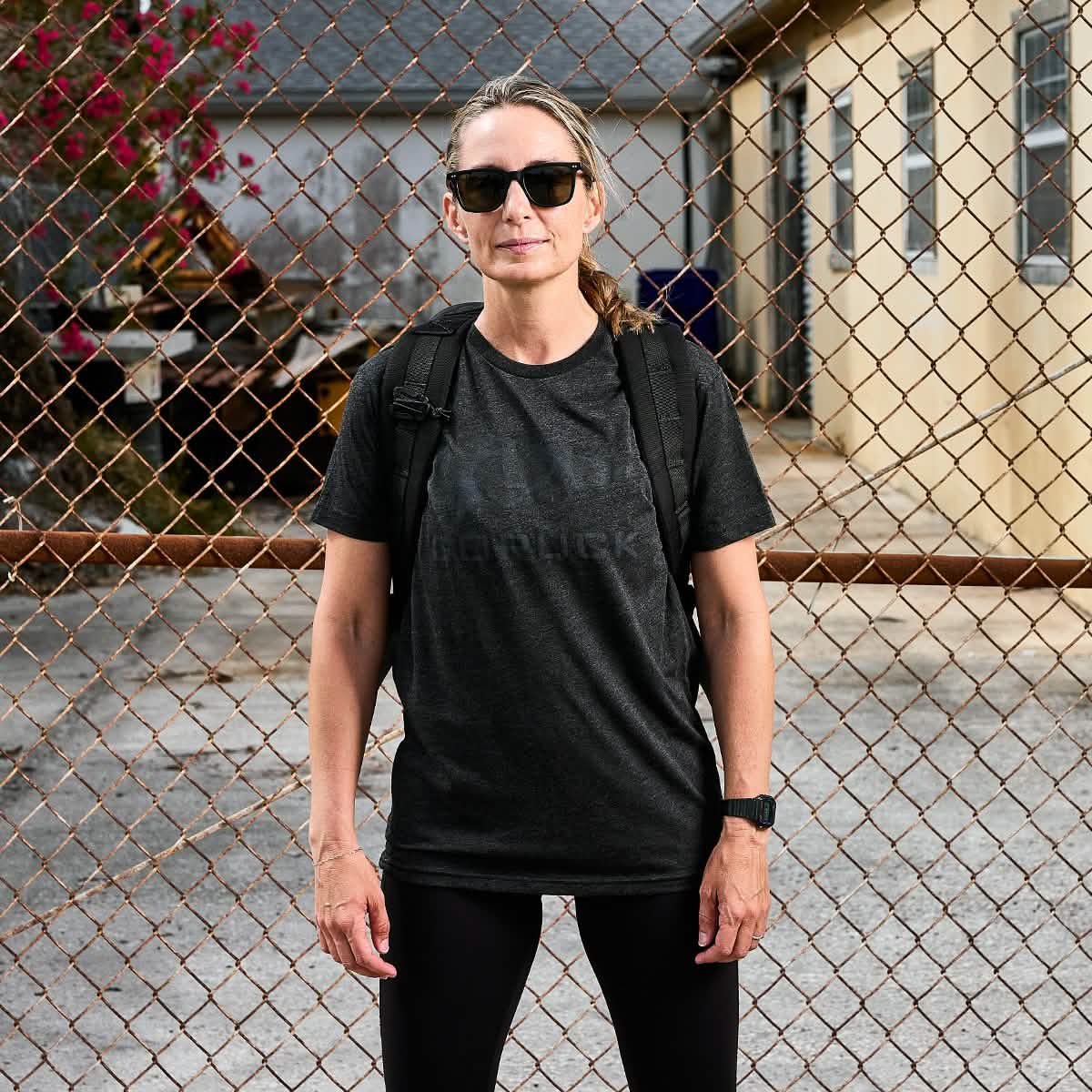 A person wearing GORUCK Spearhead sunglasses, a black GORUCK Spearhead TeeāTri-Blend from vendor-unknown, and a backpack stands in front of a chain-link fence. Premium tri-blend houses and a small tree frame the background on a cloudy day.