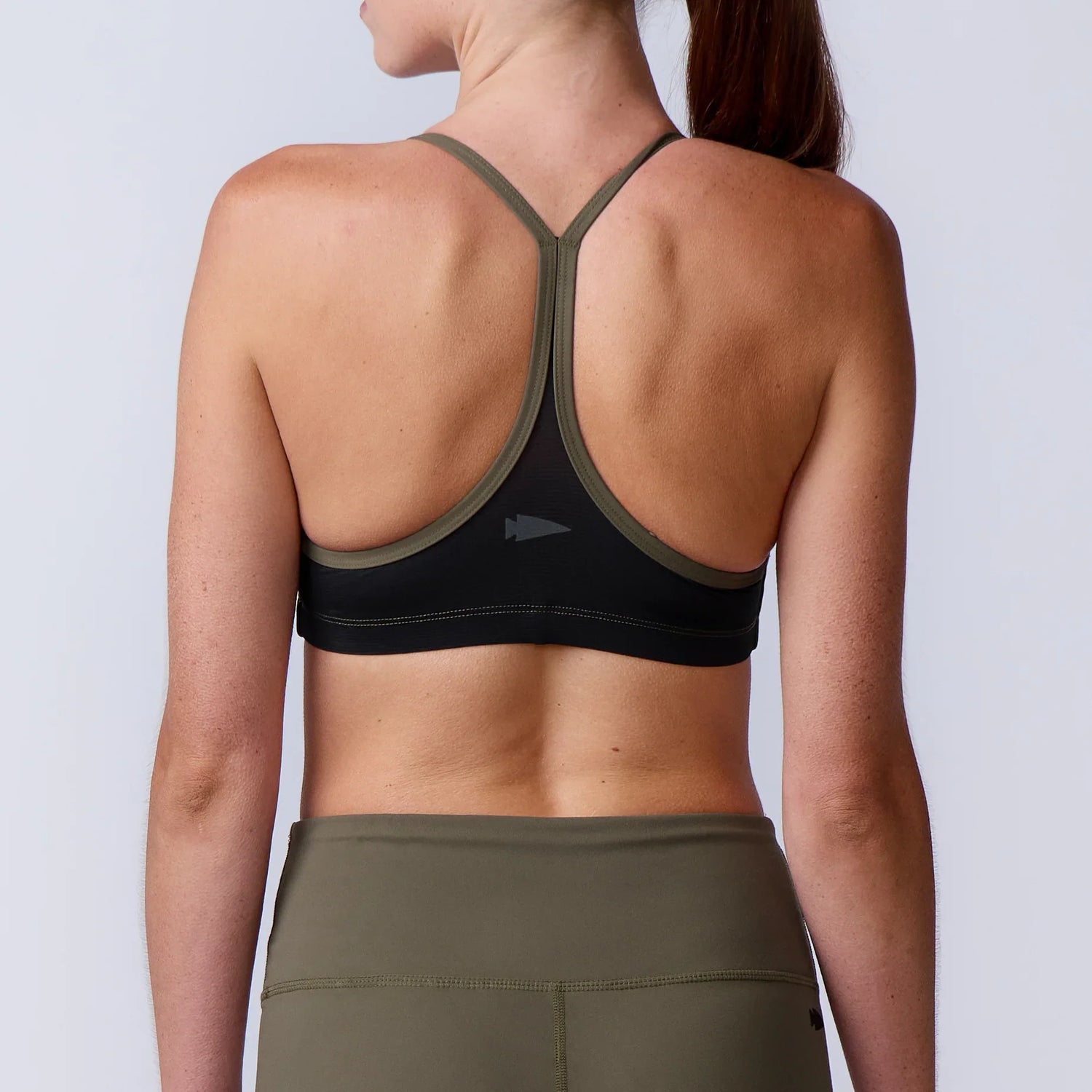 Back view of a woman wearing a black and olive green racerback sports bra and olive green leggings against a plain light background