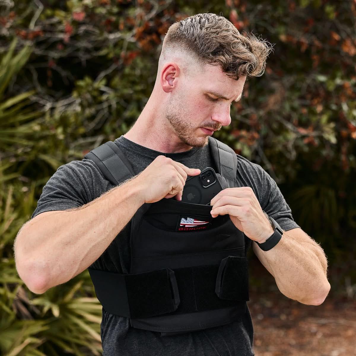 Man placing phone into front pocket of black tactical GORUCK vest outdoors with greenery background