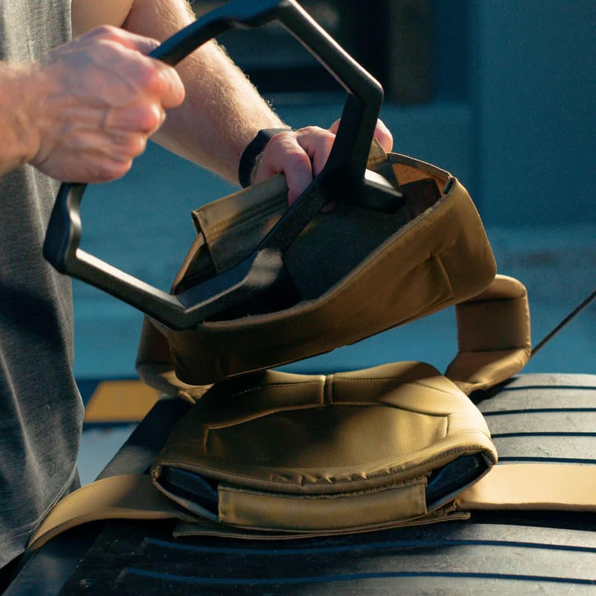 Person loading black weighted plates into tan GORUCK weighted rucksack pack outdoors