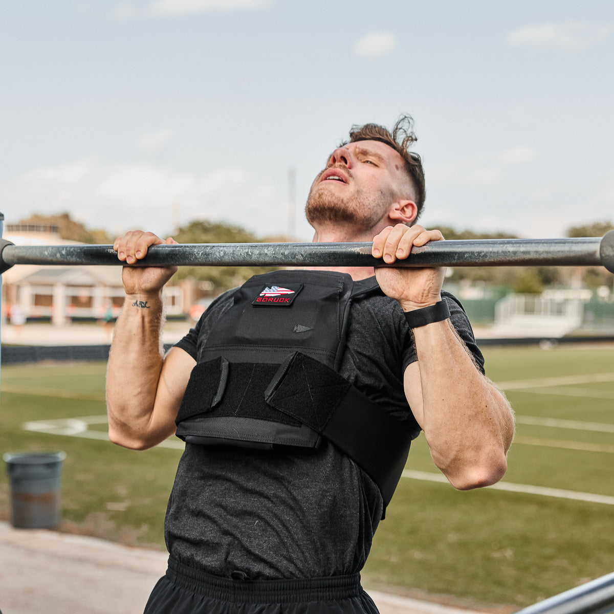 A man wearing a Rucking Weight Vest with curved plates struggles to do a pull-up on an outdoor bar at a sports field.