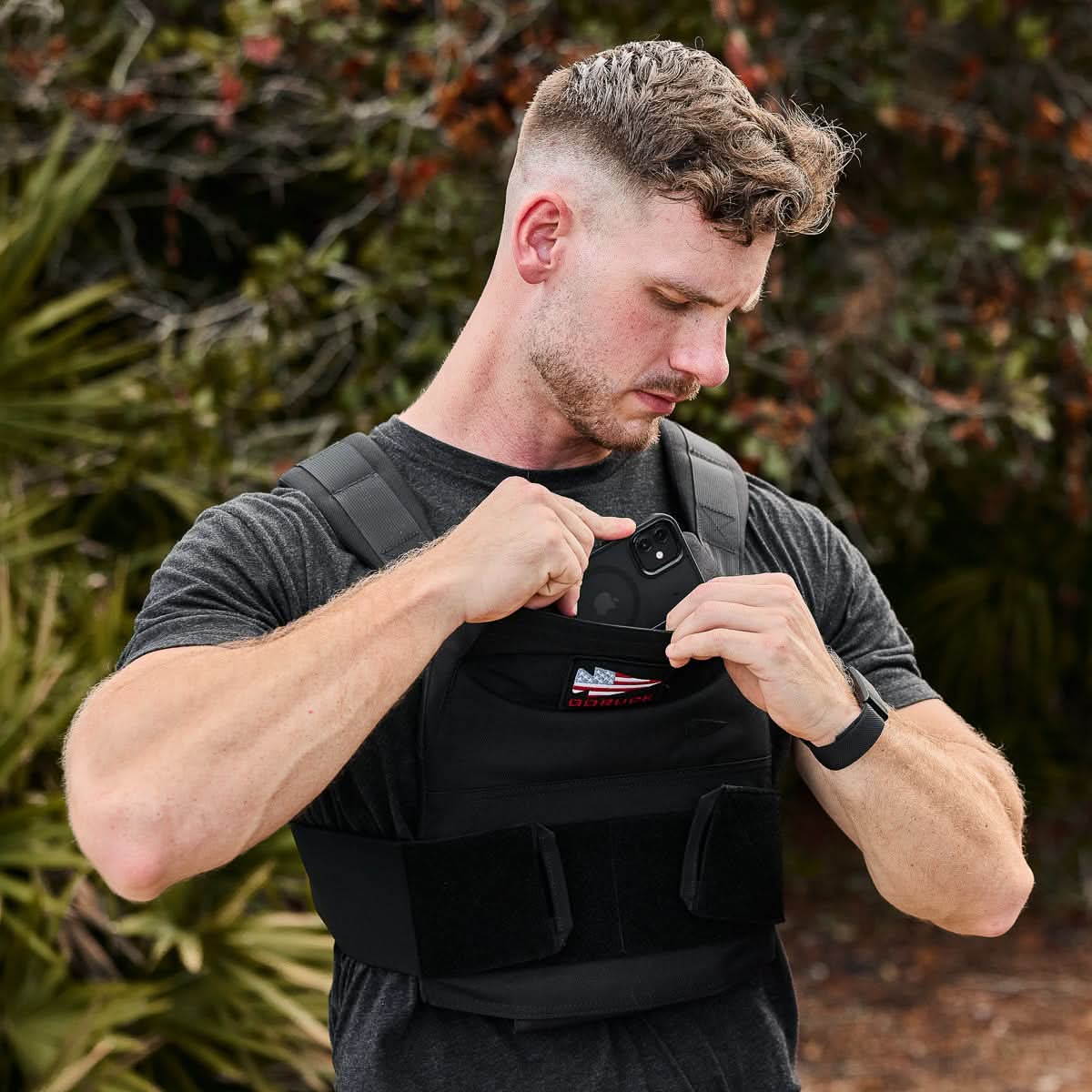 A man places his phone into the chest pocket of a Rucking Weight Vest outdoors, perfect for rucking, with trees in the background.