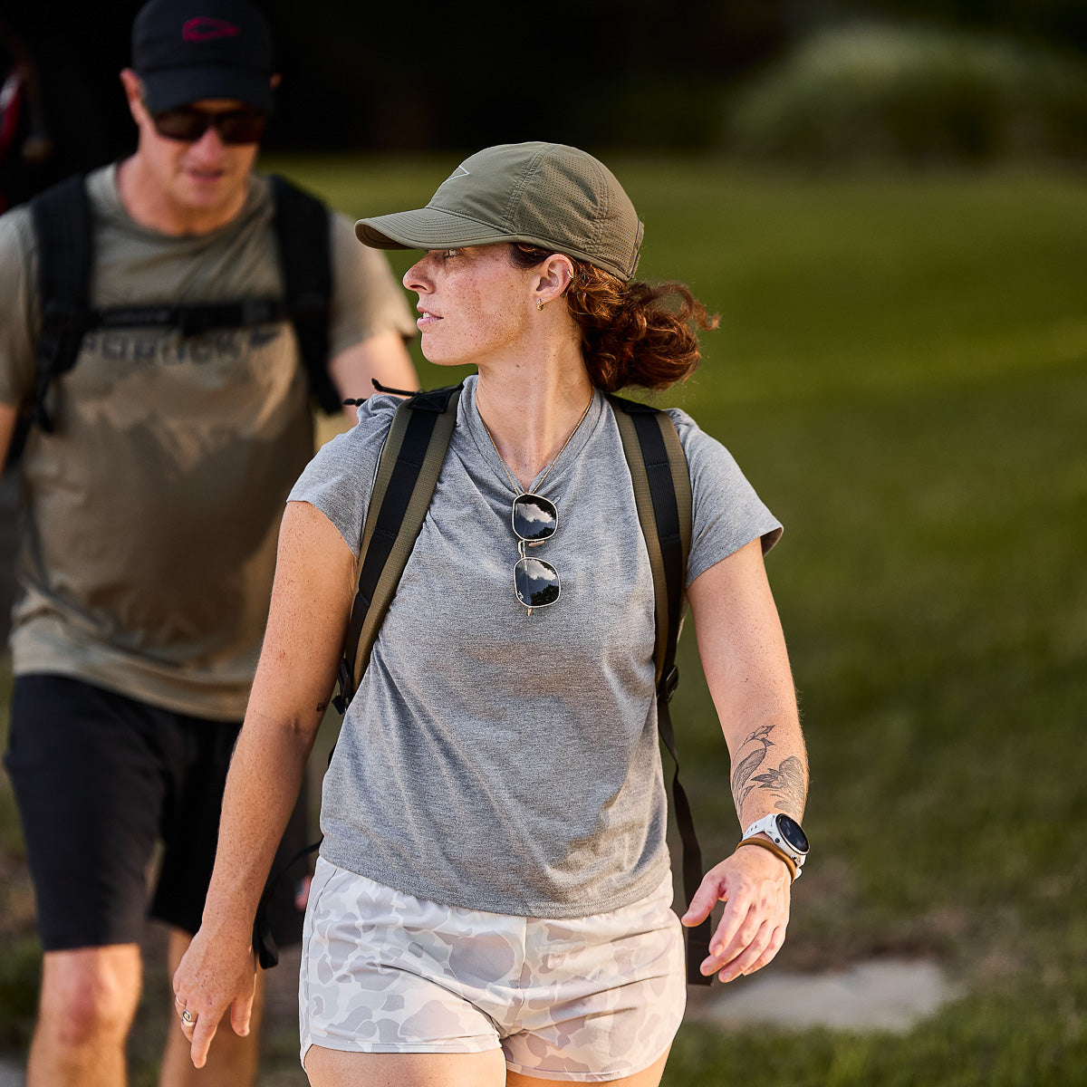 A woman in a gray shirt and cap walks outdoors with the Basic Rucker®, while a man in the background also carries a weighted rucksack—both ready for adventure.