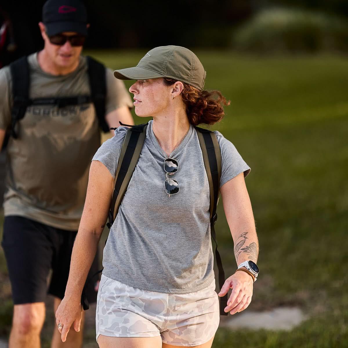 Woman wearing olive cap, gray t-shirt, and camo shorts with backpack hiking outdoors with man