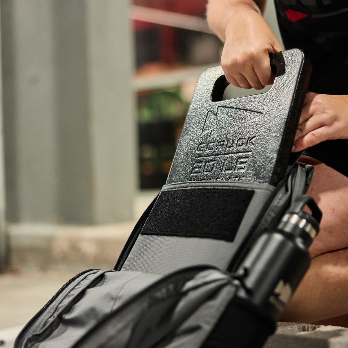 A person places a GORUCK 20 LB weight plate into their Basic Rucker® and adds a water bottle on the side, prepping a weighted rucksack for their next adventure.