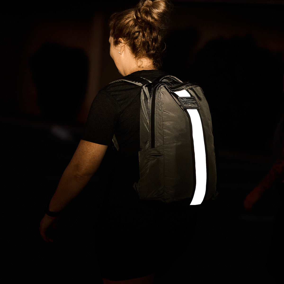 A person with a bun hairstyle wears a dark Basic Rucker® backpack with a reflective strip down the center.
