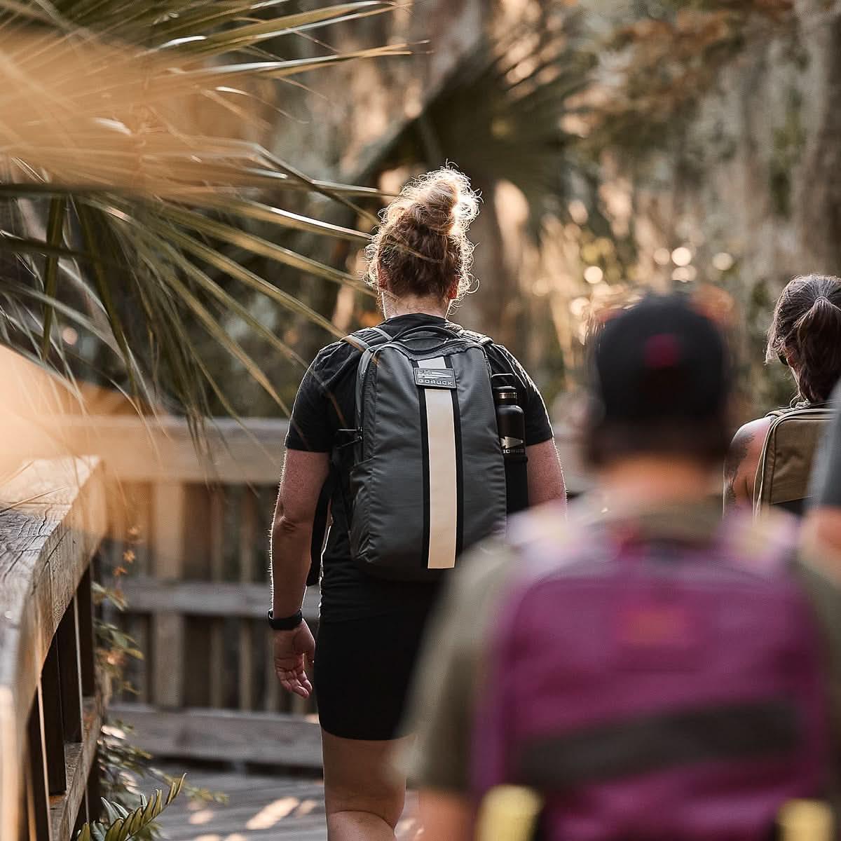 Person with a black GORUCK backpack walking on a wooden trail surrounded by natural foliage