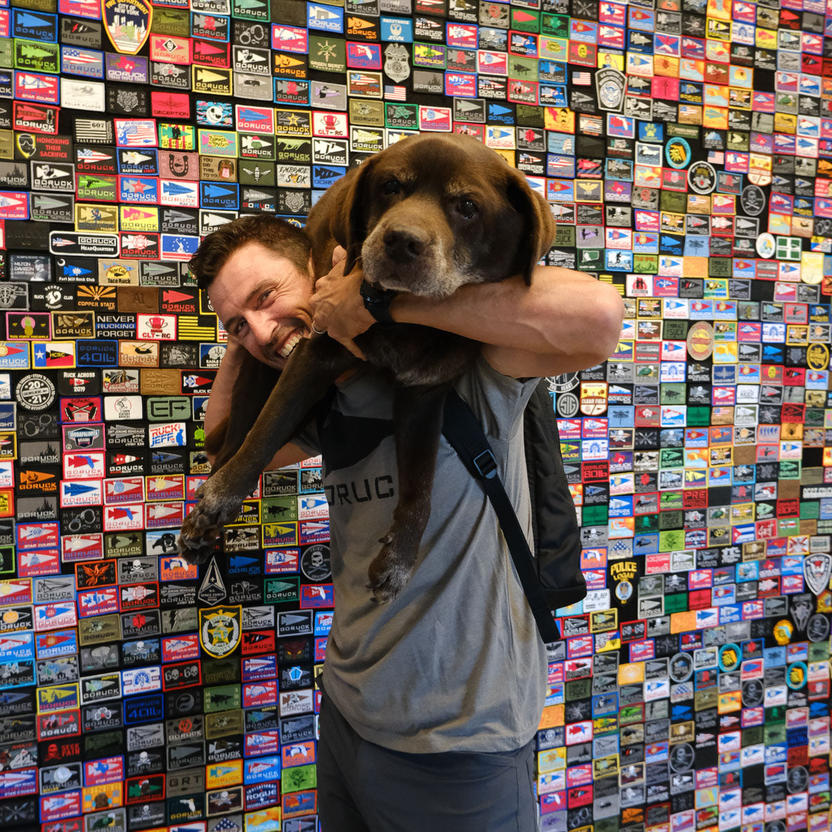 Smiling man carries a large brown dog on his shoulders in front of a colorful patch-covered wall.