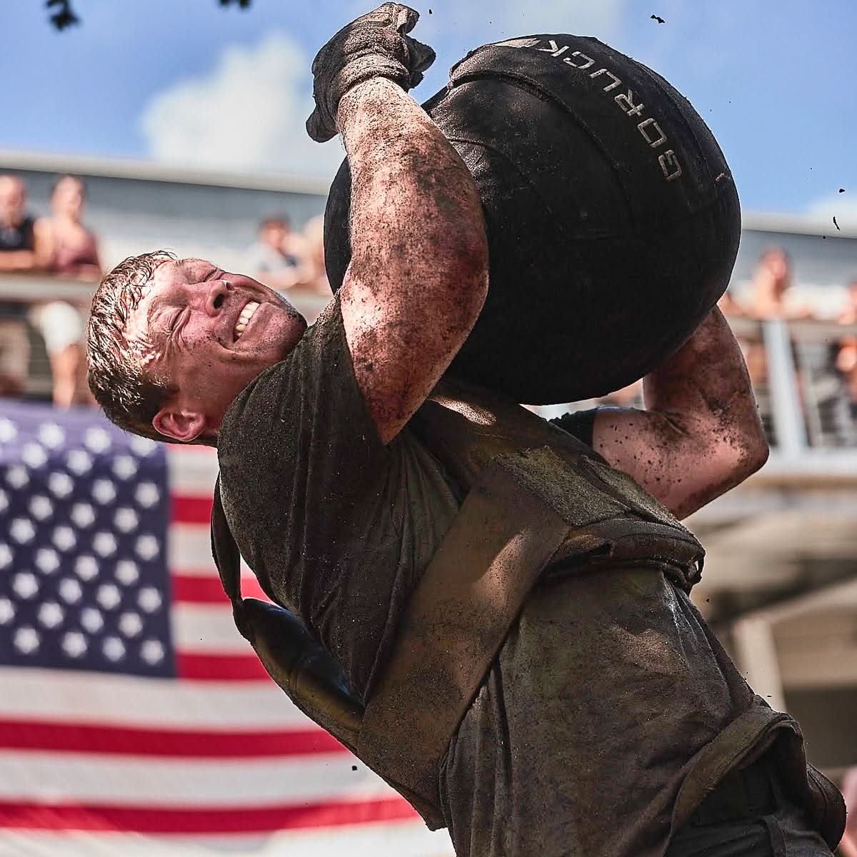 A man lifts a heavy sandbag outdoors, straining in front of a large American flag while wearing a weight vest loaded with Curved Ruck Plates for extra resistance.