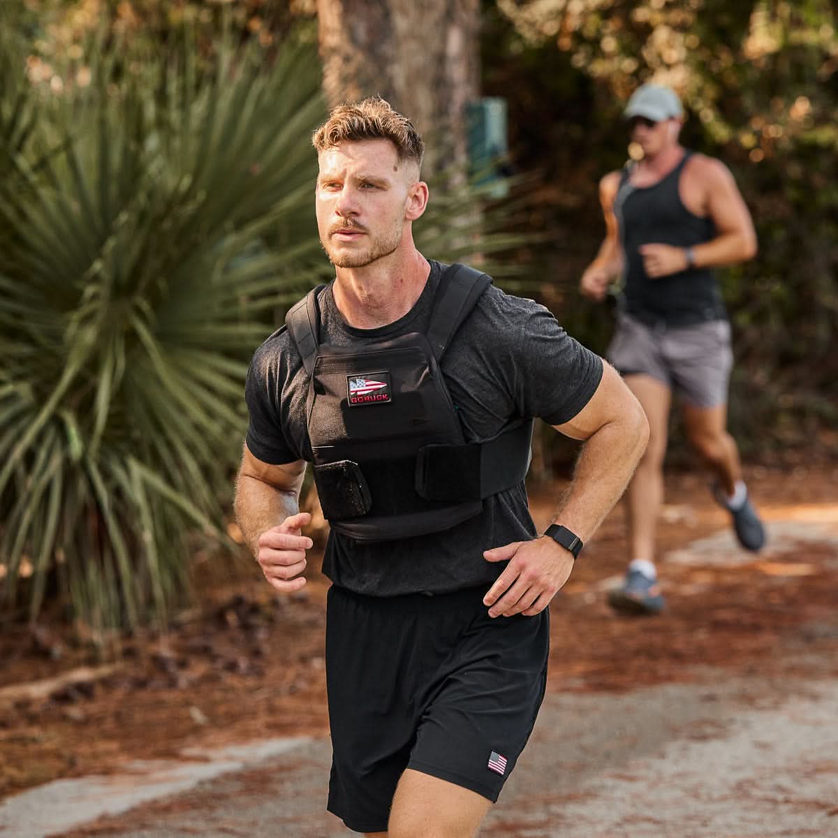 A man runs on a wooded trail wearing a Rucking Weight Vest with Curved Ruck Plates, while another runner follows behind, both embracing the intensity of rucking.