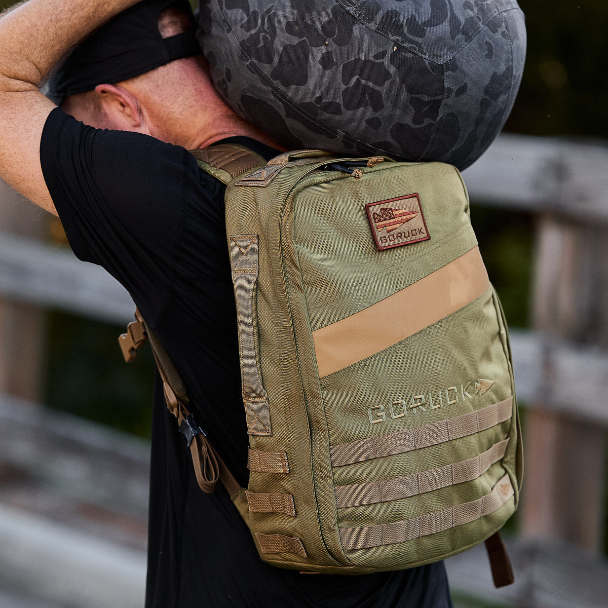Person carrying a gray camo duffel bag and wearing the olive green Rucker 4.0 outdoors, perfect for adding ruck plates during weighted training pack activities.