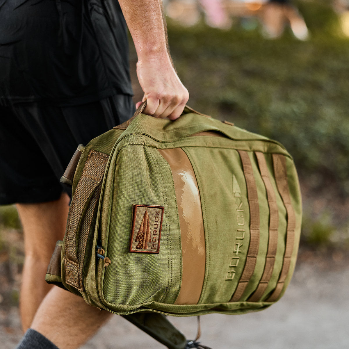 Someone outdoors carries the Rucker 4.0, an olive green rucking backpack, by its top handle while wearing black shorts.