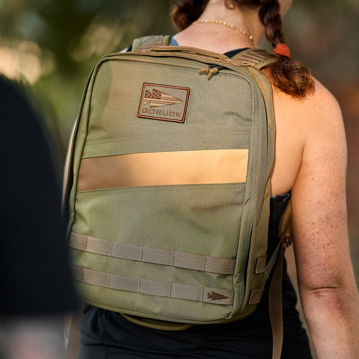 A person outdoors, seen from behind, wears an olive green Rucker 4.0 backpack with ruck plate compatibility as sunlight shines on the bag.
