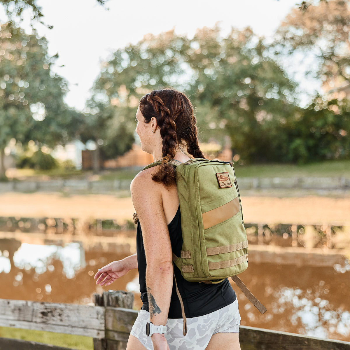 A woman with braids, wearing a green Rucker 4.0 backpack, walks outdoors by a river on a sunny day.