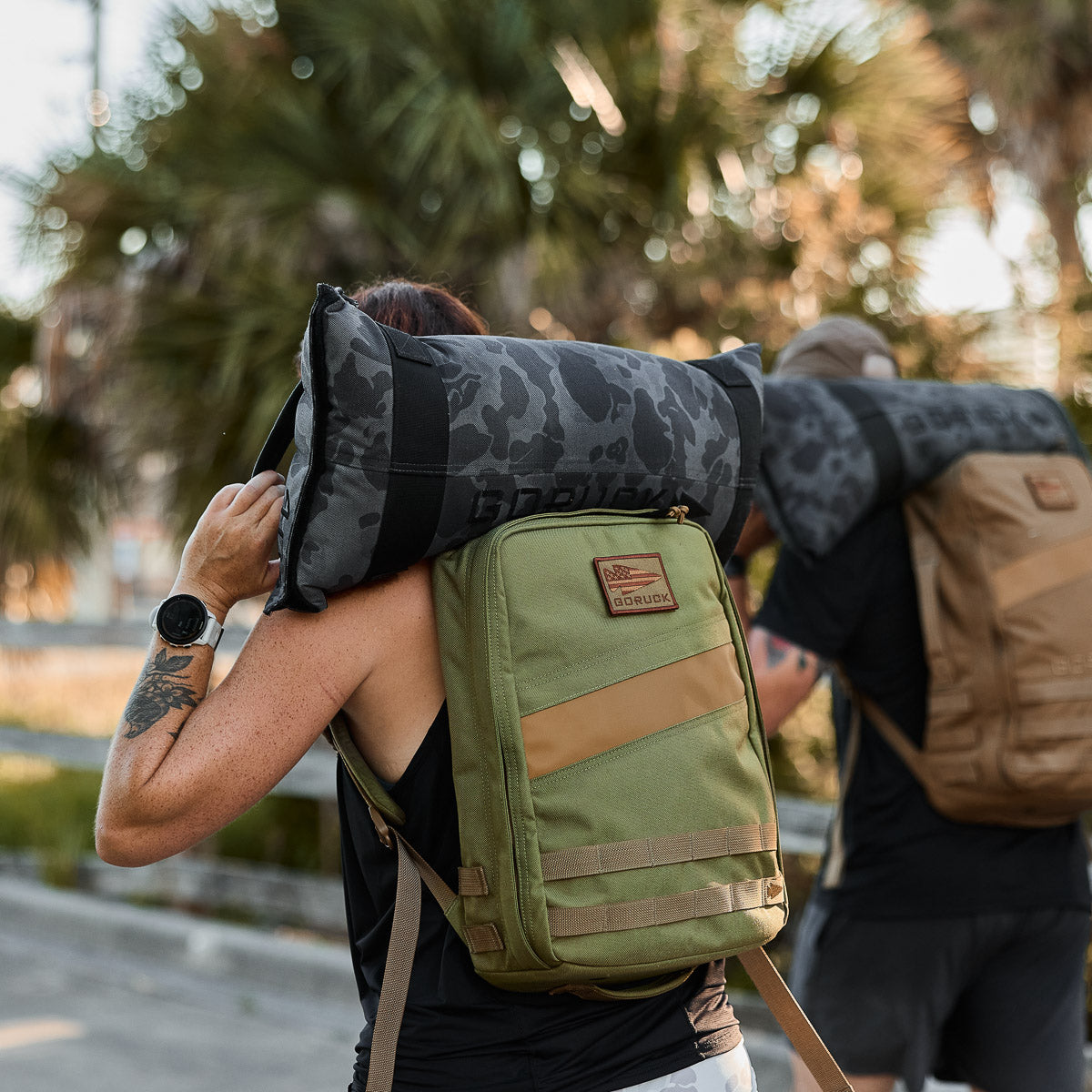 Two people carry Rucker 4.0 backpacks and camo-patterned sandbags on their shoulders outdoors by palm trees.