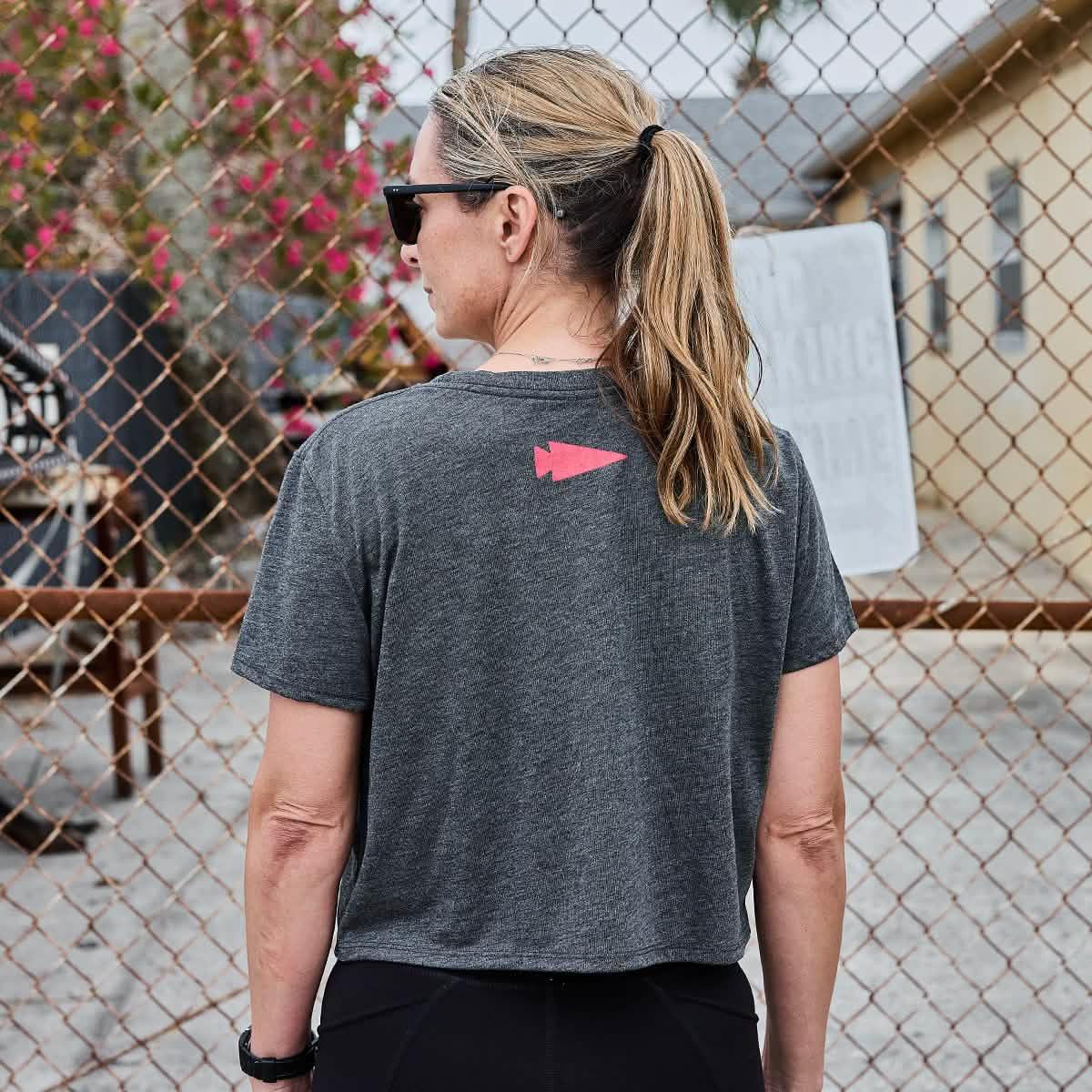 Woman wearing gray GORUCK t-shirt with red logo on back, standing near chain-link fence outdoors