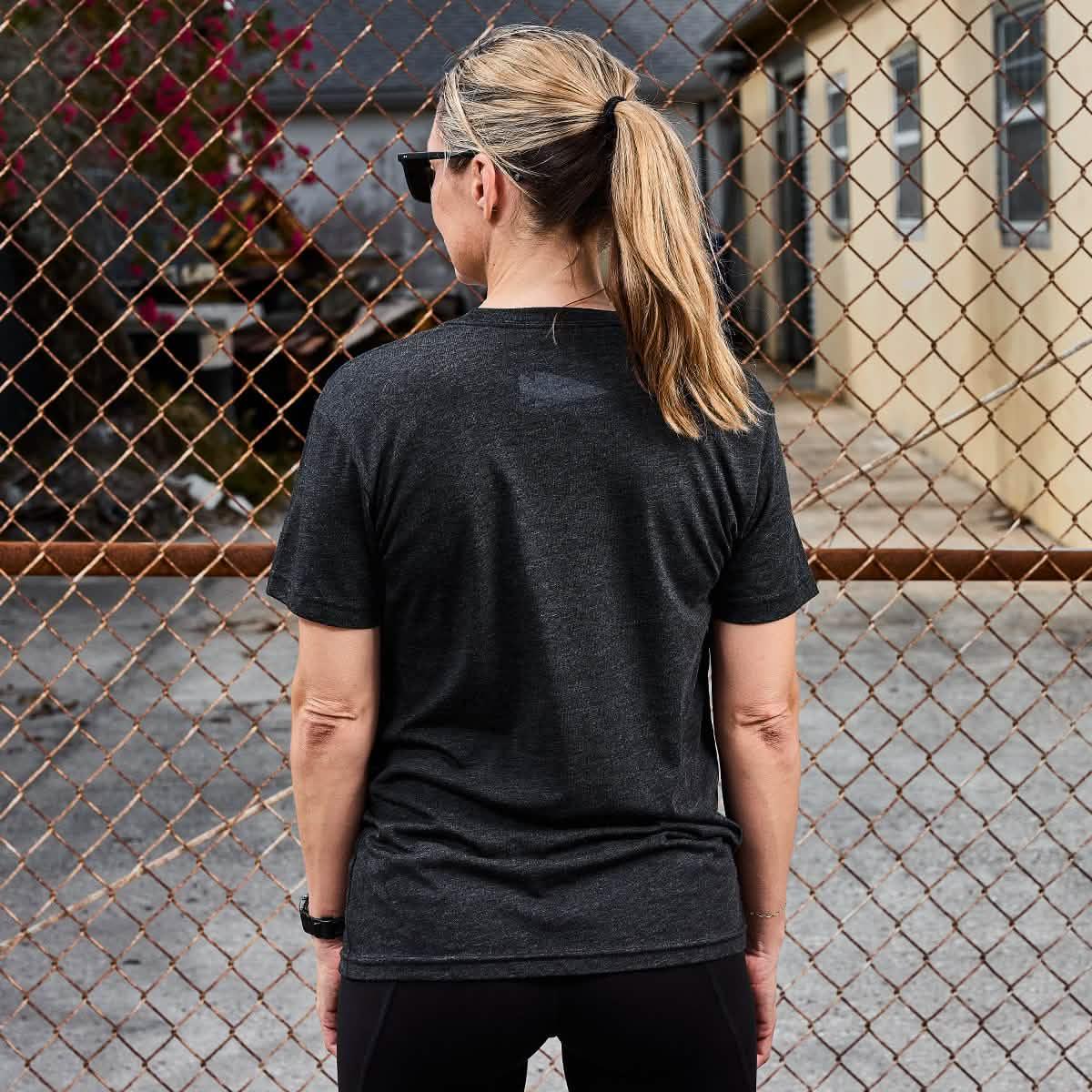 Woman with blonde ponytail wearing black sunglasses and dark grey t-shirt standing facing chain-link fence outdoors