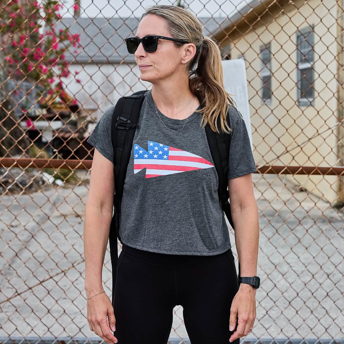 A person wearing sunglasses and a GORUCK Women's Cropped Tee - Poly-Blend, featuring a stylized American flag design, stands in front of a chain-link fence. With a backpack slung over one shoulder, they appear to be outdoors on a cloudy day, perhaps after an invigorating workout.