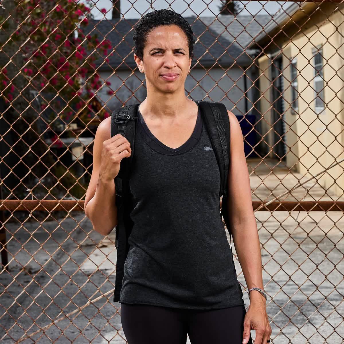 Person wearing a black sleeveless rucking shirt and backpack standing in front of a chain-link fence outdoors