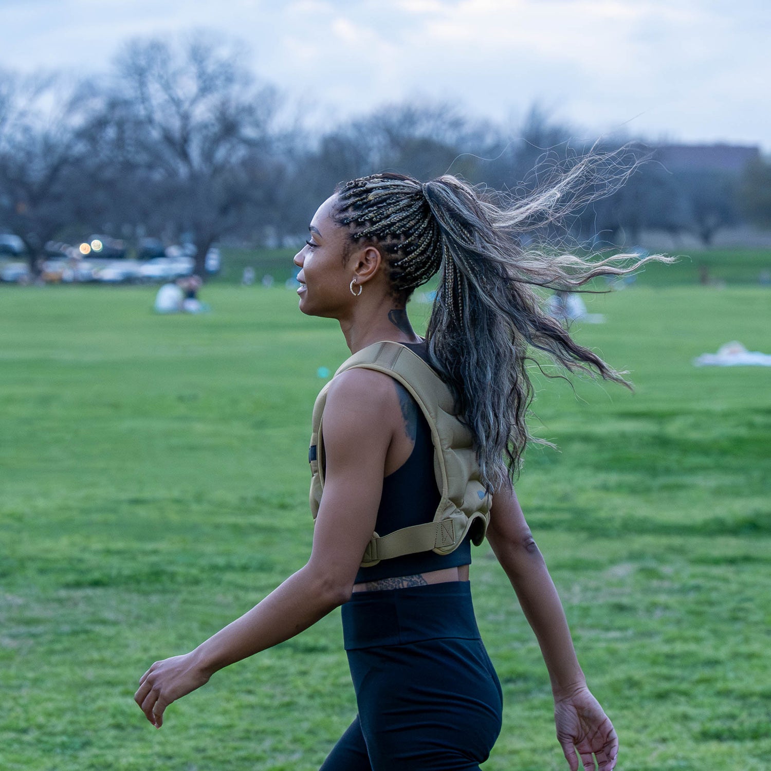 A woman with braided hair wears the Spy Ruck | Women's Weighted Vest while walking outdoors on green grass, with trees and people in the background.