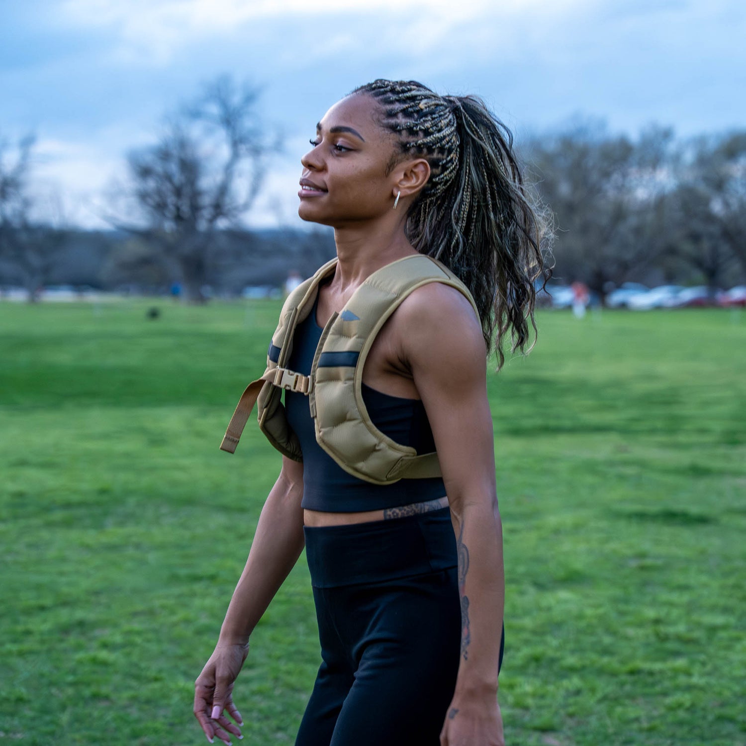 A woman with braided hair, dressed in athletic wear, walks outdoors on a grassy field wearing the Spy Ruck | Women's Weighted Vest.