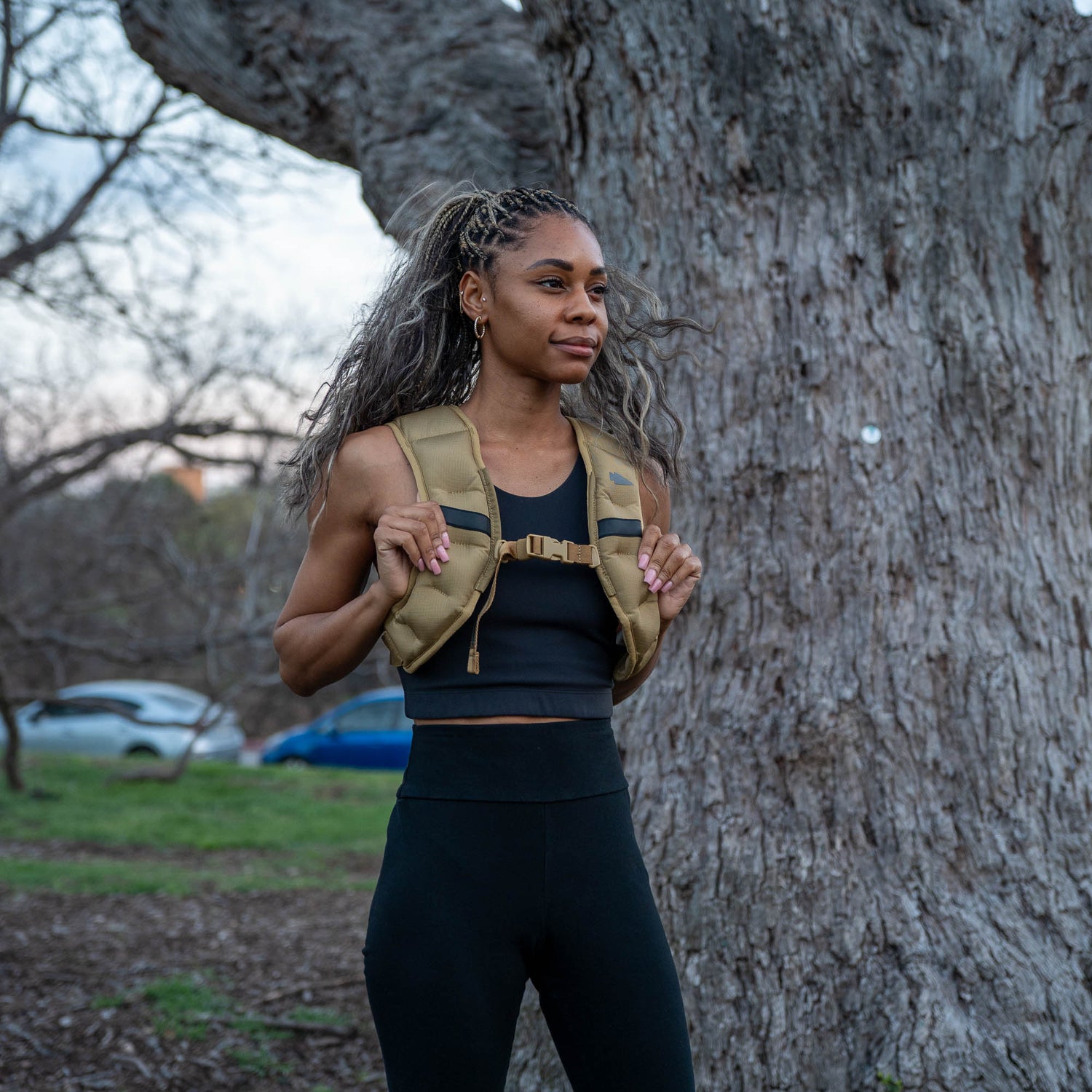 A woman in athletic wear stands outdoors in front of a large tree, wearing the Spy Ruck | Women's Weighted Vest.