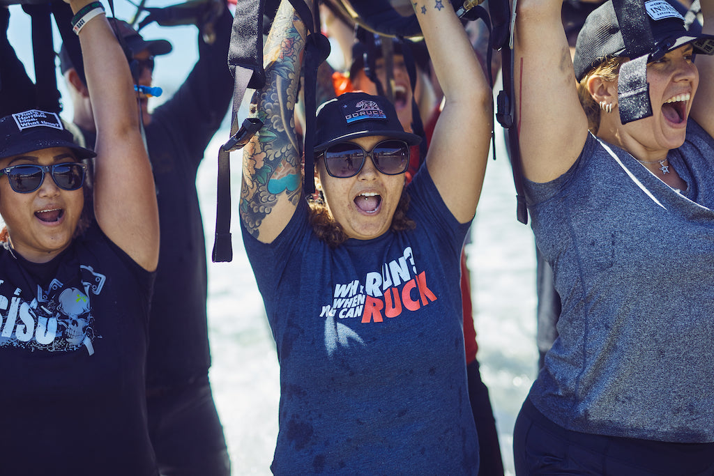 A group of women outdoors cheer with arms raised, wearing hats and sunglasses, looking excited and energetic.