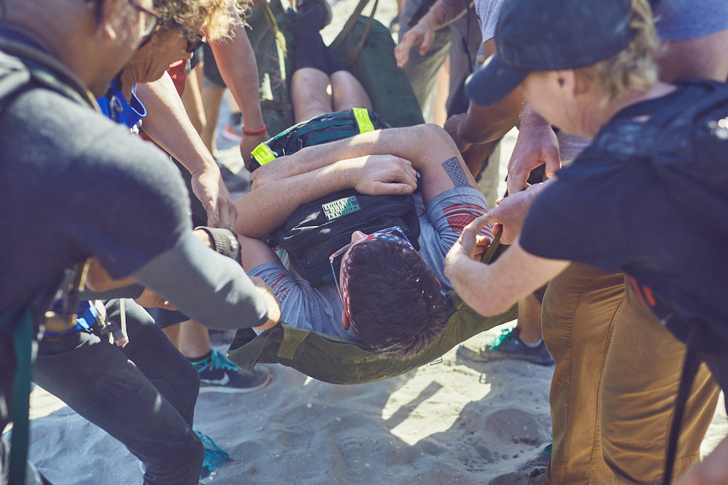 Several people carry a person on a stretcher across sandy ground, collaborating in an emergency situation.