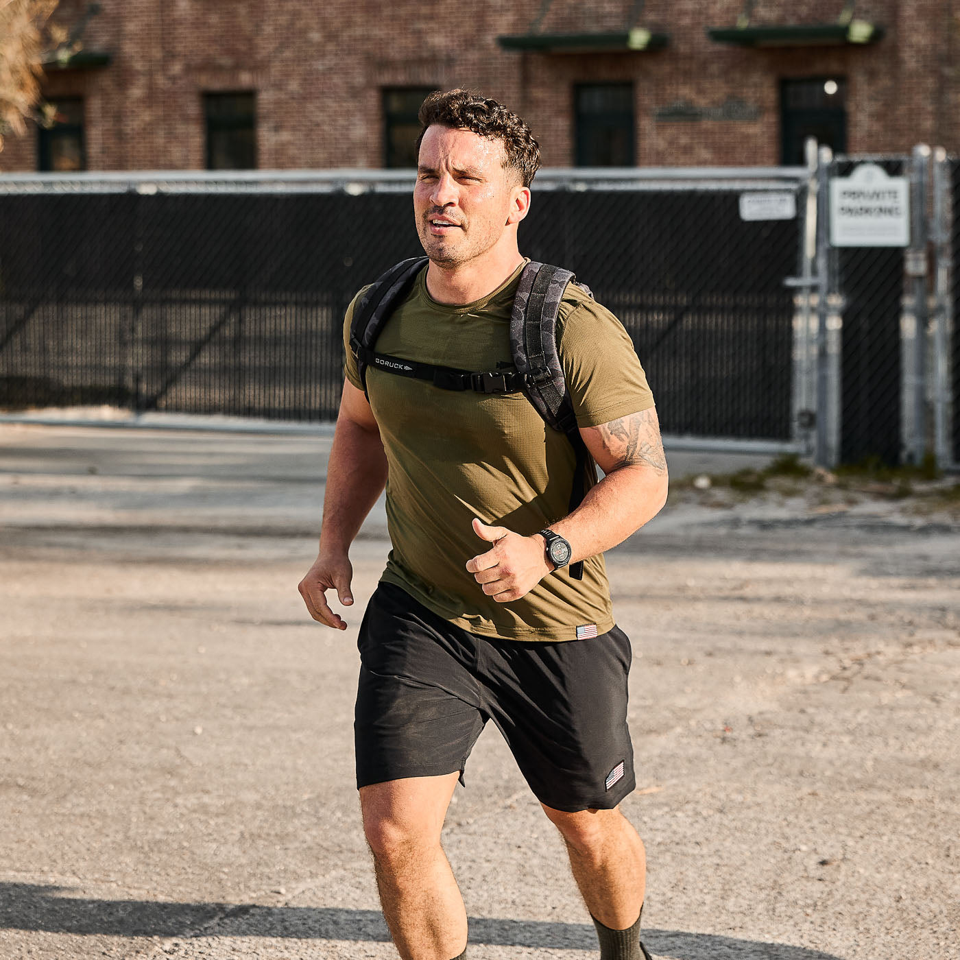 A man wearing the Ruck Plate Carrier 3.0 with ergonomic lumbar support runs outdoors on a sunny day, passing a brick building and fence in the background.