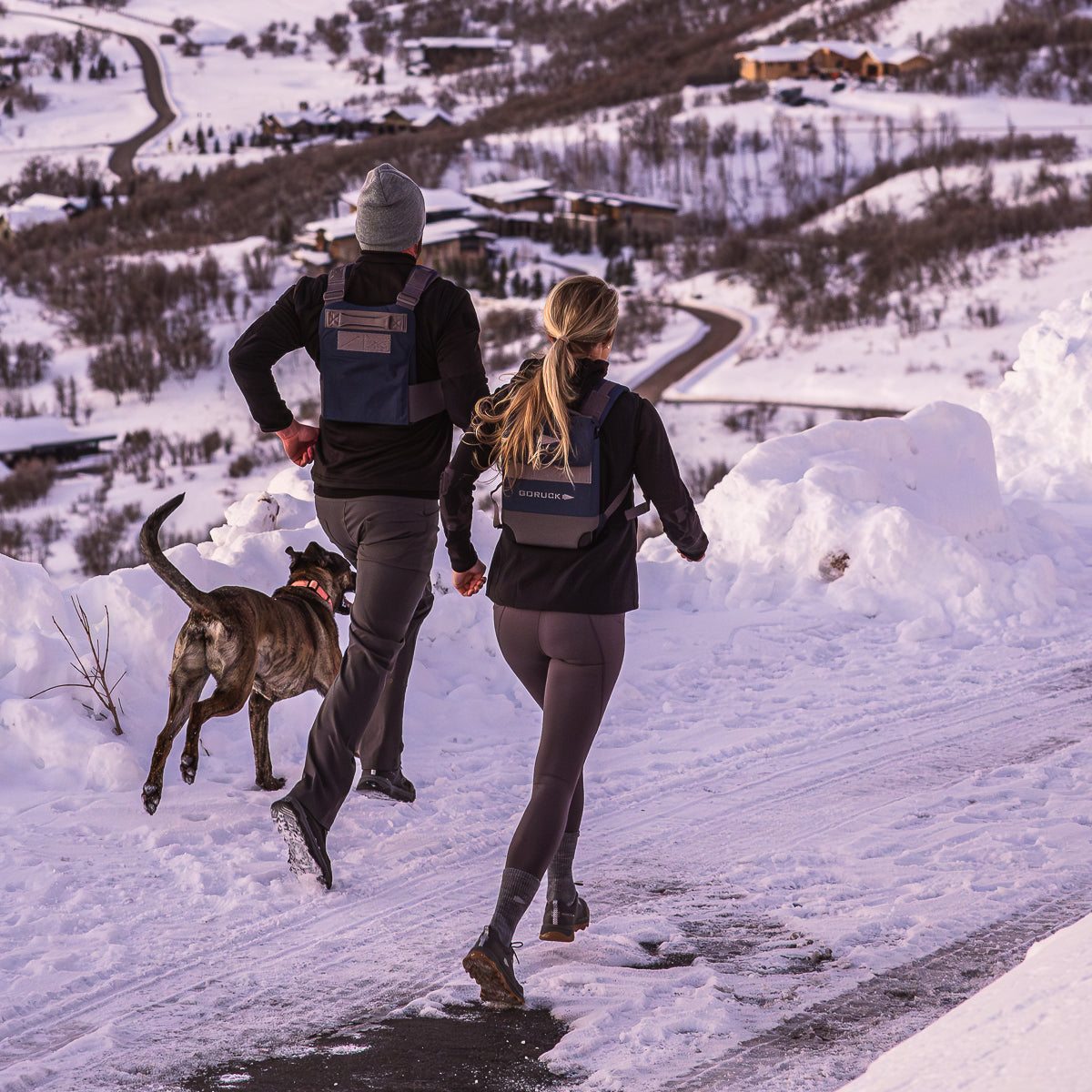 Two people and a dog run on a snowy hill overlooking a winding road and trees. One wears the ergonomic Ruck Plate Carrier 3.0, making their winter adventure comfortable with padded shoulder straps allowing free movement in the crisp mountain air.