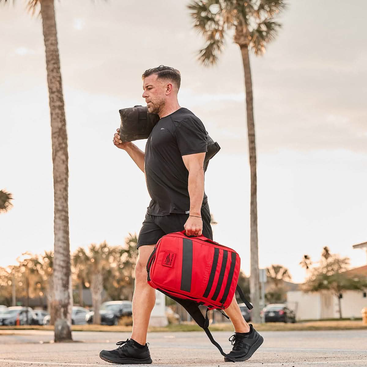 Clad in GORUCK's Men's Rough Runner - Blackout athletic wear and lightweight running shoes, an individual walks outdoors. They carry a red and black backpack in one hand and have a black duffel bag slung over their shoulder as they stride past palm trees and parked cars under a cloudy sky, seemingly ready for the challenge.