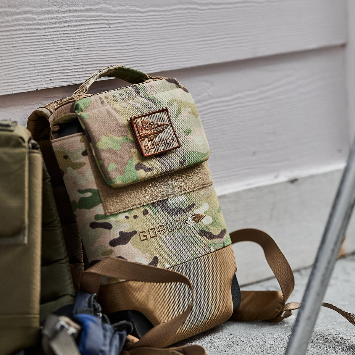 The Ruck Plate Carrier 3.0, a camouflage GORUCK backpack with tan padded shoulder straps and a velcro patch, rests on concrete near a white wall, blending comfort and style.