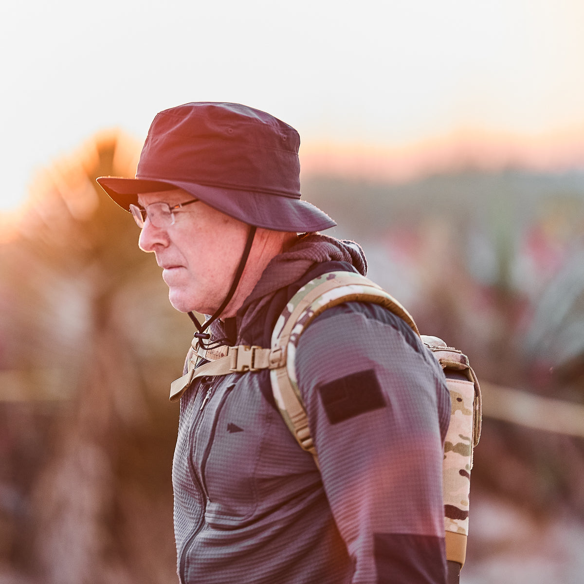 A man wearing glasses, a black hat, and the Ruck Plate Carrier 3.0 backpack with padded shoulder straps and ergonomic lumbar support walks outdoors at sunrise or sunset.