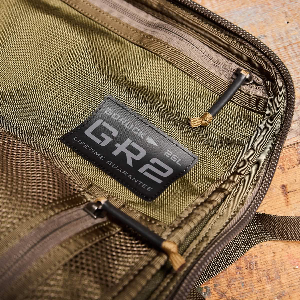 Close-up of an olive green rucksack labeled GORUCK GR2 - Cordura, featuring a Lifetime Guarantee. The image showcases the zipper details and stitching, emphasizing its durability and quality inspired by Special Forces. The bag is partially unzipped, revealing its interior.