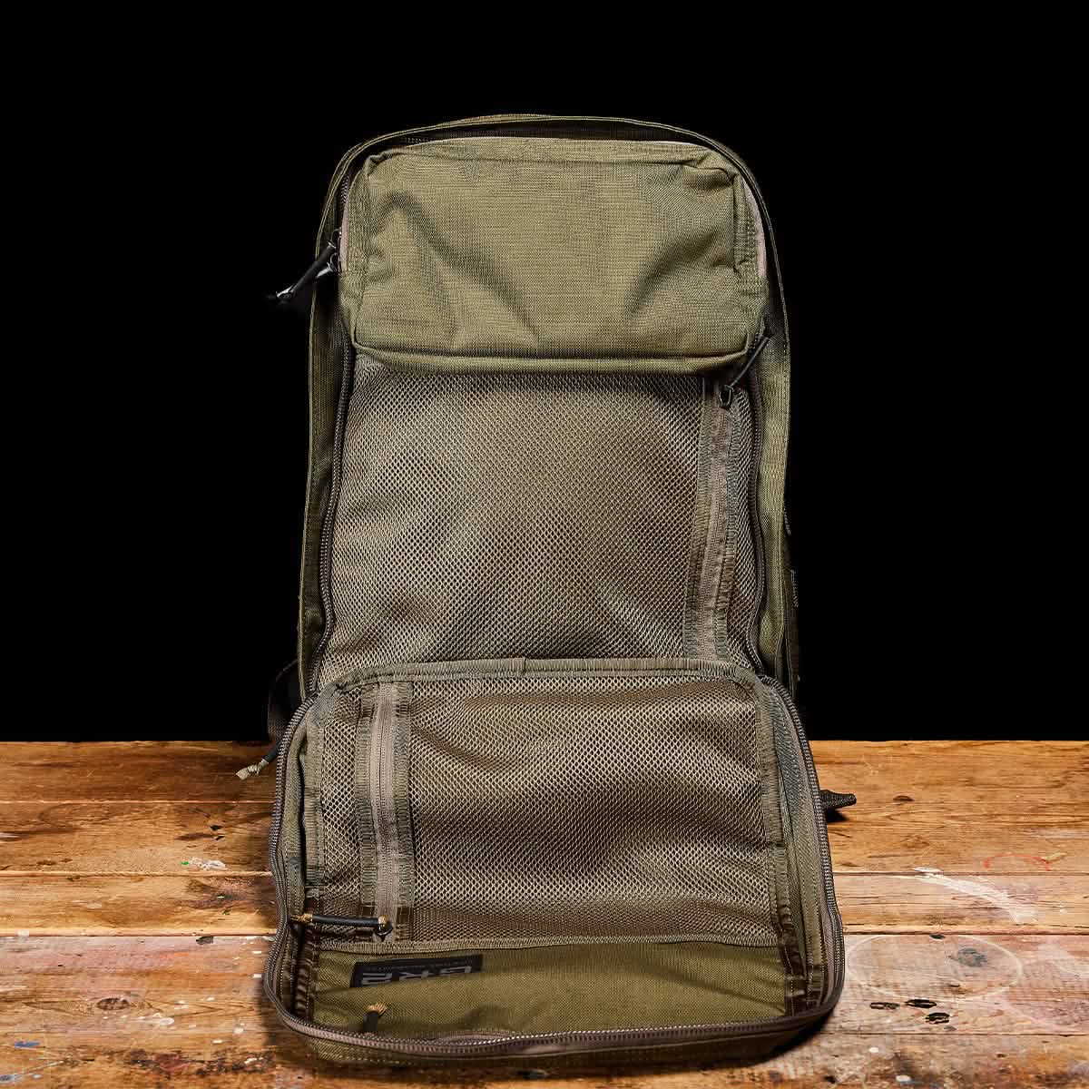 A GORUCK GR2 - Cordura backpack, in olive-green and evoking the style of a Special Forces rucksack, is shown open on a wooden surface against a black background. It includes multiple mesh compartments and pockets for efficient organization. The interior is visible, highlighting its roomy GR2-inspired design.