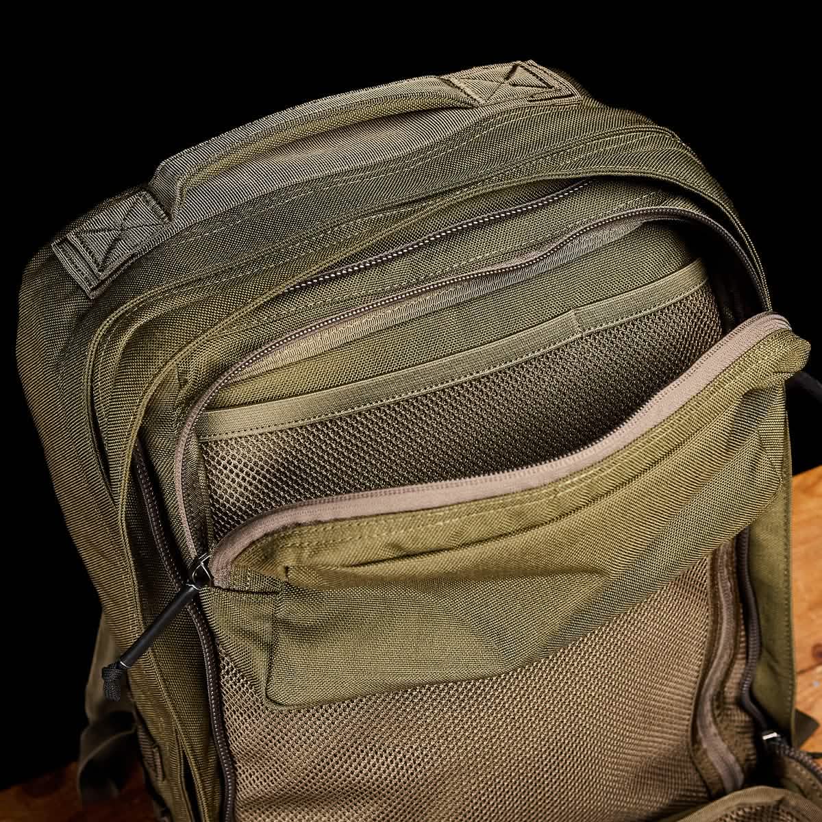 A close-up of a green GR2 - Cordura by GORUCK highlights its numerous zippered compartments. The ruck is open, displaying mesh-lined pockets, set against a dark background on a wooden surface, evoking an aura of Special Forces efficiency.