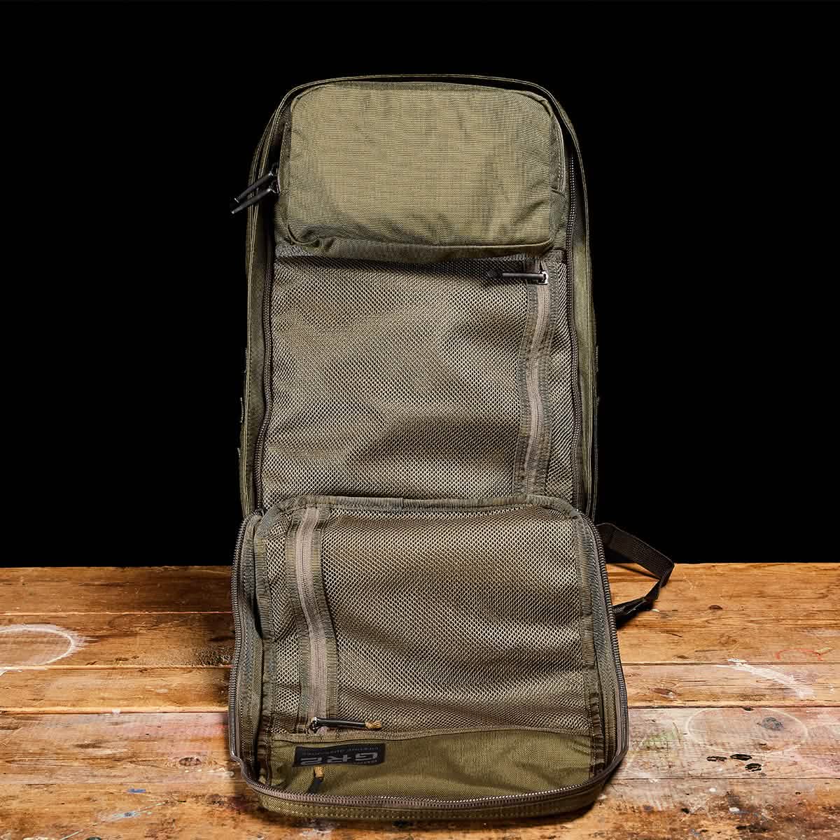 The GR2 by GORUCK, an olive green travel bag inspired by those used by Special Forces, stands upright on a wooden surface against a black background. Featuring multiple zippered compartments and mesh pockets, it offers organized storage space ideal for any adventure.