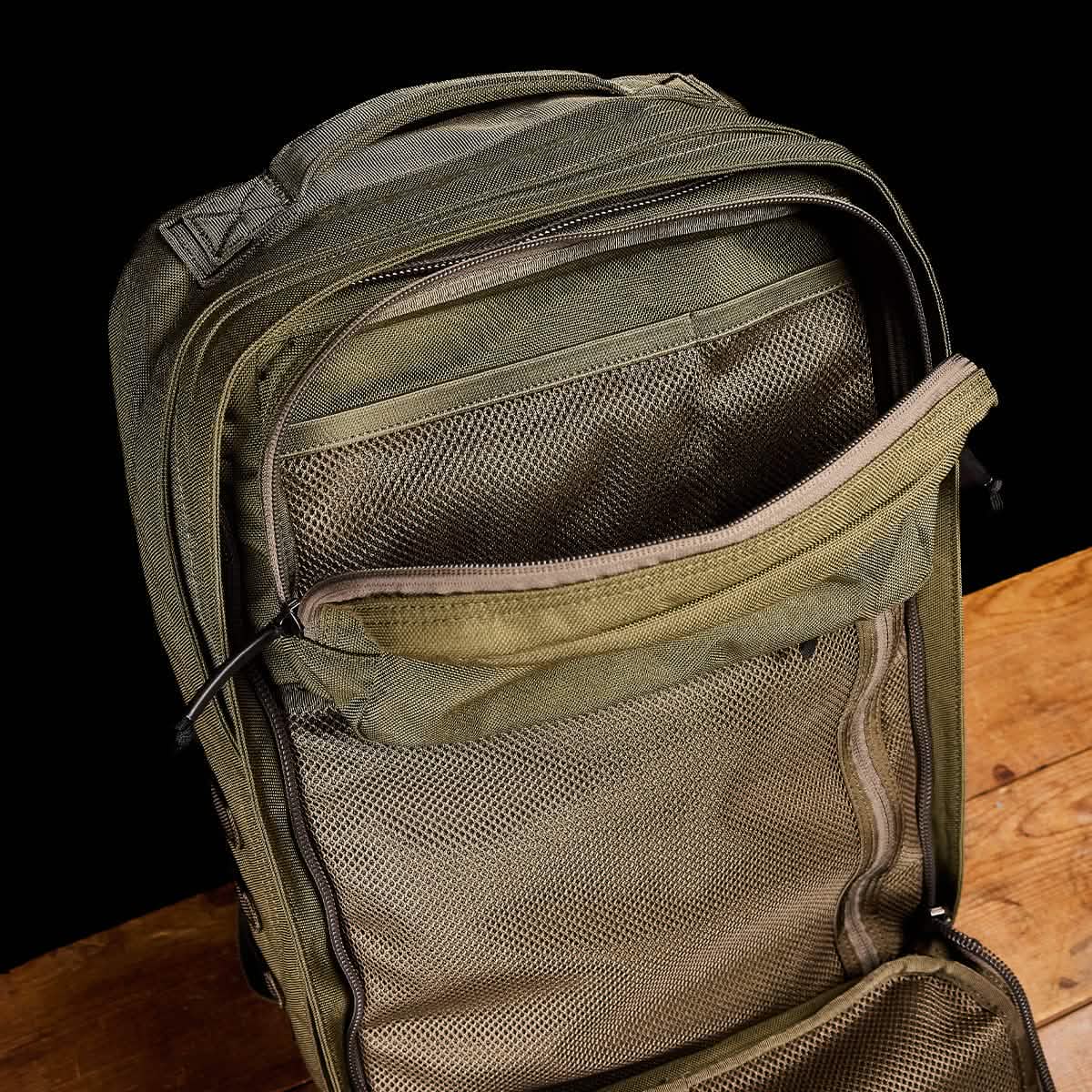 The open GR2 backpack from GORUCK, in green, sits on a wooden surface against a black backdrop. Its Special Forces quality is clear as the unzipped main compartment displays mesh pockets for exceptional organization, making it perfect for those looking for carry-on compliant options.