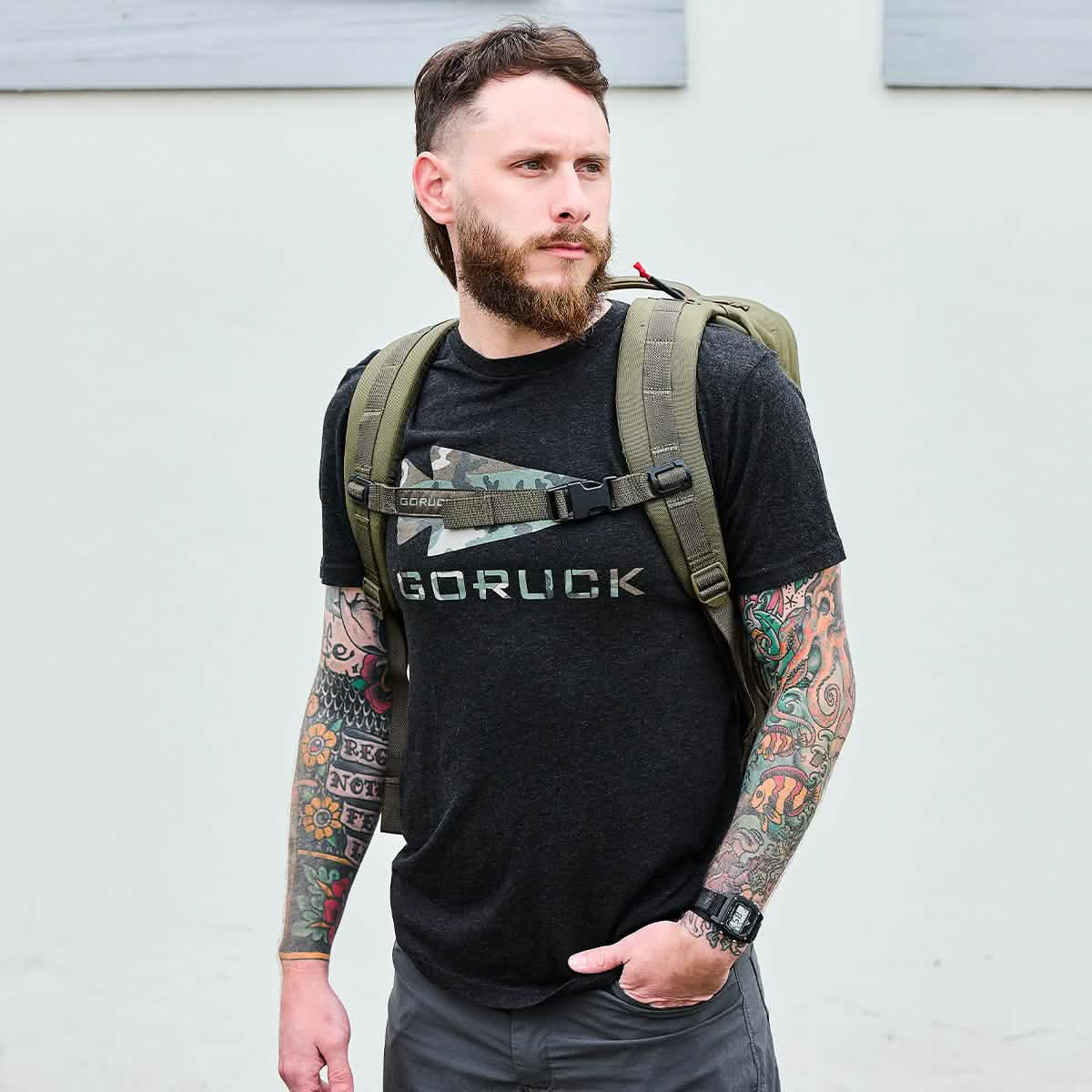 Bearded man with tattoos wearing a black GORUCK t-shirt and olive green rucksack against a plain light background