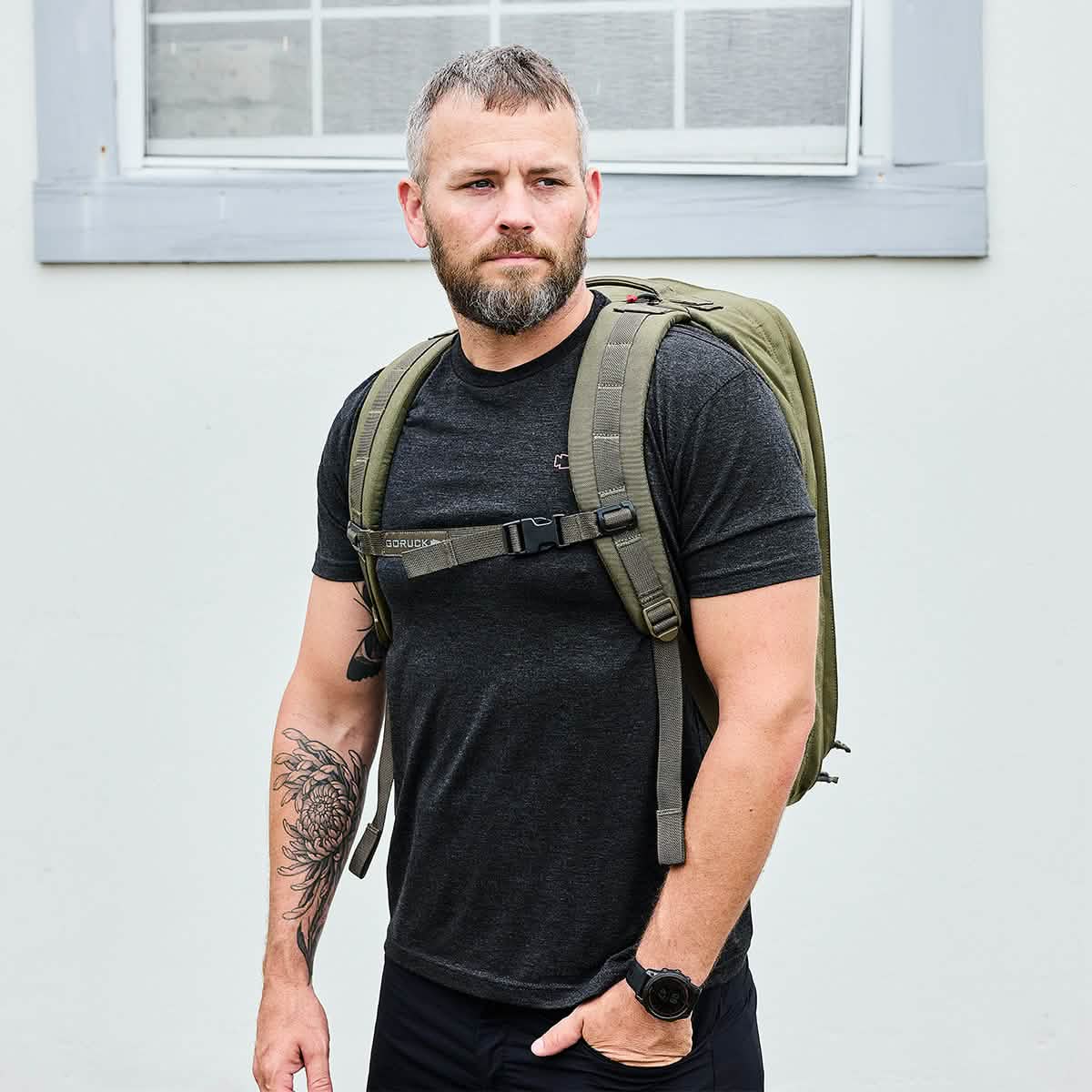 A man with short hair and a beard stands outside wearing a black t-shirt and carrying a GORUCK GR2 - Cordura ruck. He has a tattoo on his right arm and wears a watch on his left wrist. The background features a light-colored wall with a window.