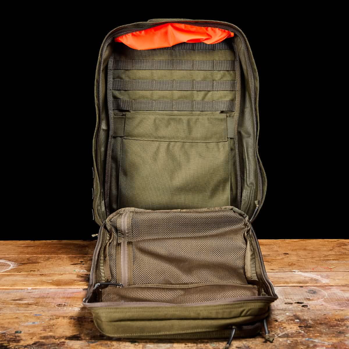 A GR2 - Cordura by GORUCK, a green tactical backpack resembling those used by Special Forces, is displayed on a wooden surface with its orange interior visible. Inside, it features multiple mesh compartments and webbing for organization. The ruck stands empty and upright against a black background.