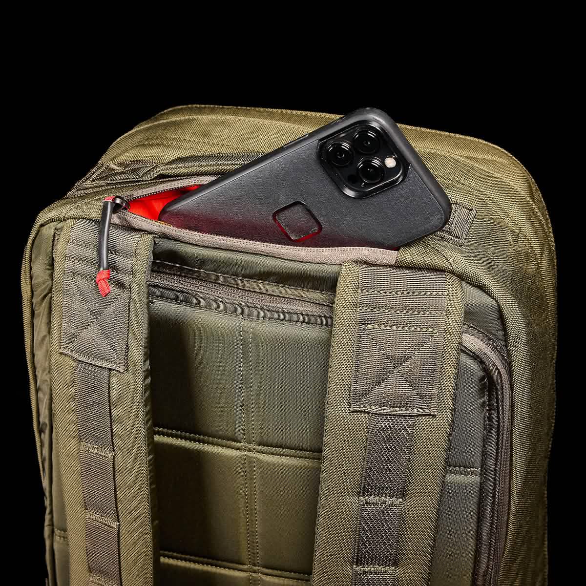 The GR2 - Cordura by GORUCK, a black smartphone rests in the partially open exterior pocket of a green, textured ruck against a black background; red accents highlight the zipper pull and inside the pocket, reminiscent of Special Forces precision.