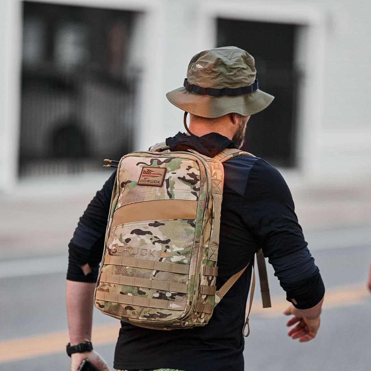 Person wearing GORUCK multi-cam backpack and ranger green boonie hat walking outdoors on urban street