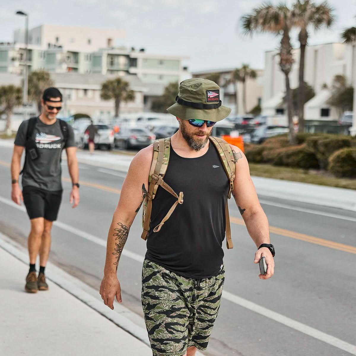 Man wearing a ranger green GORUCK boonie hat, black tank top, camo shorts, and backpack walking on a street near palm trees